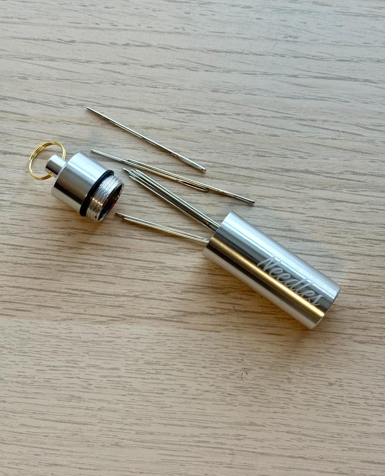 May include: A cylindrical silver metal needle case with a screw-top lid and a gold-colored keyring. The word Needles is engraved on the side of the case. Several thin, metallic sewing needles are positioned inside and outside of the open container.