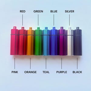May include: A row of ten cylindrical metal pill containers with loops at the top. The colors are labeled as pink, red, orange, green, teal, blue, purple, silver, and black. Each container features a screw-top lid and a smooth, metallic finish.