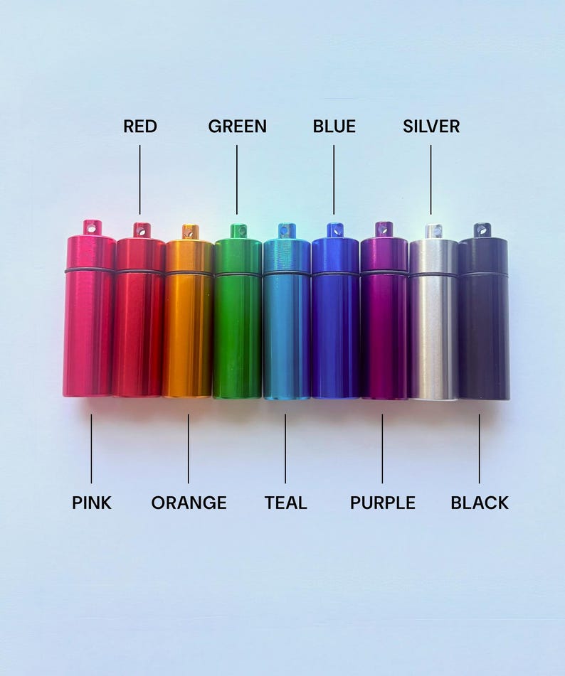 May include: A row of ten cylindrical metal pill containers with loops at the top. The colors are labeled as pink, red, orange, green, teal, blue, purple, silver, and black. Each container features a screw-top lid and a smooth, metallic finish.