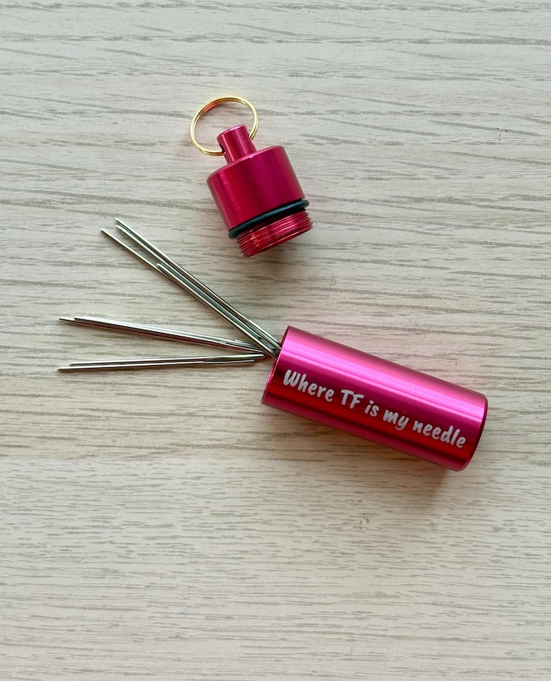 May include: A cylindrical magenta metal needle case with a screw-top lid and a gold-tone keyring. The case features the white text "Where TF is my needle" printed on the side. Several thin silver sewing needles protrude from the open container.