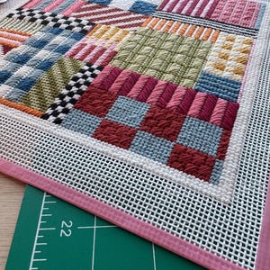 May include: A colorful needlepoint project with a variety of stitched patterns and textures. The design features squares and rectangles in shades of red, blue, pink, green, and white. The needlepoint is framed with a white mesh and a pink border.