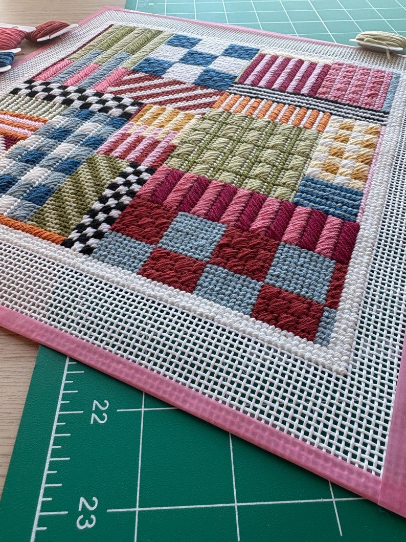 May include: A colorful needlepoint project with a variety of stitched patterns and textures. The design features squares and rectangles in shades of red, blue, pink, green, and white. The needlepoint is framed with a white mesh and a pink border.