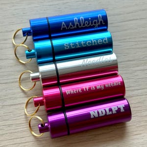 May include: Five cylindrical metal needle cases with gold-toned key rings. The cases are blue, light blue, silver, pink, and purple. They feature laser-engraved text: Ashleigh, Stitched, Needles, where TF is my needle, and NDLPT.