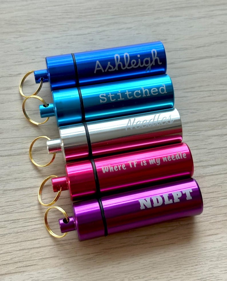 May include: Five cylindrical metal needle cases with gold-toned key rings. The cases are blue, light blue, silver, pink, and purple. They feature laser-engraved text: Ashleigh, Stitched, Needles, where TF is my needle, and NDLPT.