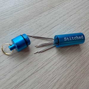 May include: A bright blue cylindrical metal needle case with a screw-top lid and a gold-toned keyring. The word Stitched is printed in white on the side of the case. Several thin silver sewing needles are protruding from the open container.