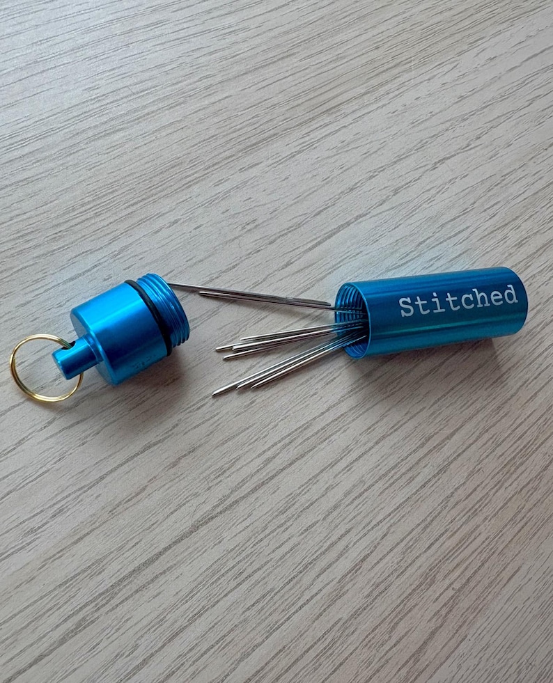 May include: A bright blue cylindrical metal needle case with a screw-top lid and a gold-toned keyring. The word Stitched is printed in white on the side of the case. Several thin silver sewing needles are protruding from the open container.