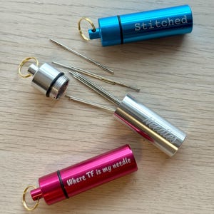 May include: Three cylindrical metal needle cases with key rings. One blue case is engraved with "Stitched", one silver case is engraved with "Needles" and contains several sewing needles, and one pink case is engraved with "Where TF is my needle".
