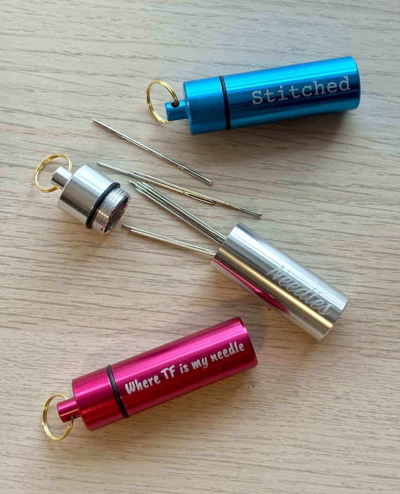 May include: Three cylindrical metal needle cases with key rings. One blue case is engraved with "Stitched", one silver case is engraved with "Needles" and contains several sewing needles, and one pink case is engraved with "Where TF is my needle".