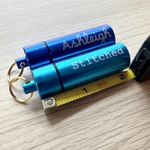 May include: Two cylindrical metal pill cases with key rings, one royal blue engraved with the name Ashleigh and one turquoise engraved with the word Stitched. A yellow measuring tape shows the cases are approximately 2.25 inches in length.