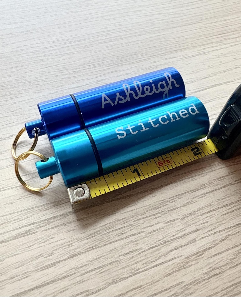 May include: Two cylindrical metal pill cases with key rings, one royal blue engraved with the name Ashleigh and one turquoise engraved with the word Stitched. A yellow measuring tape shows the cases are approximately 2.25 inches in length.