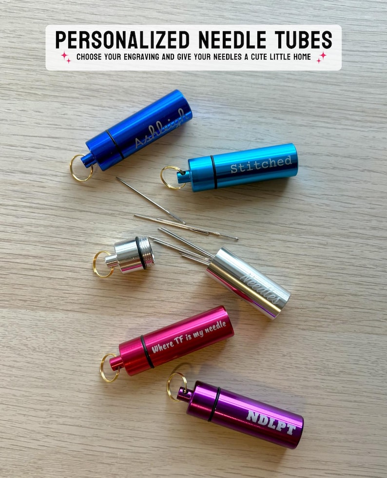 May include: Five cylindrical metal needle tubes in blue, light blue, silver, red, and purple, each with a gold keyring. The tubes feature engravings including names and phrases like 'Stitched', 'Needles', 'Where TF is my needle', and 'NDLPT'.