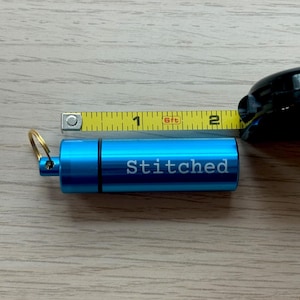 May include: A bright blue cylindrical metal container with a gold-toned keyring attached to one end. The word Stitched is engraved in white on the side. A yellow measuring tape shows the container is approximately 2 inches in total length.