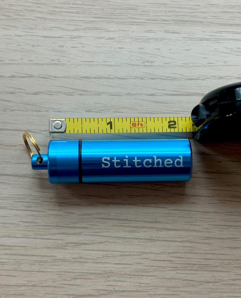May include: A bright blue cylindrical metal container with a gold-toned keyring attached to one end. The word Stitched is engraved in white on the side. A yellow measuring tape shows the container is approximately 2 inches in total length.