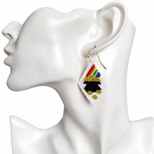 May include: A single beaded earring with a gold hook, featuring a white, diamond-shaped design. The design includes a black pot of gold with a rainbow and gold accents. The earring is displayed on a white mannequin head.
