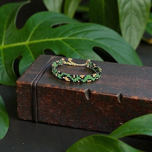 May include: A beaded bracelet with a geometric pattern in black, green, and gold is displayed on a dark brown, rectangular box. The bracelet has gold-colored clasps and a delicate chain. Large green leaves provide a natural backdrop.