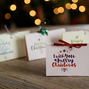 May include: A collection of Christmas gift tags with festive messages. The tags are in various colors, including pink and cream, and are adorned with red, green, and white text. A red ribbon is tied on one tag. The background features blurred Christmas lights.