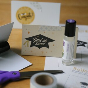 May include: A graduation-themed gift set featuring rollerball bottles, greeting cards, and stationery. The cards display graduation cap graphics and the text "You're Done!" and "Congrats!". A pair of purple scissors and tape are also visible.