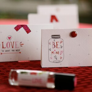 May include: Valentine's Day themed gift tags with a red and white color scheme. One tag reads "LOVE is what you need" and another says "BE mine" inside a jar illustration. A small bottle of lip gloss is in the foreground.