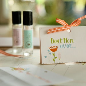 May include: A gift set featuring two glass roll-on bottles with black caps, a greeting card with the text "Best Mom ever...", and an orange and white checkered ribbon. The card has a floral design.