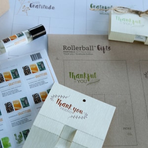 May include: An assortment of gift items, including a rollerball bottle, stickers, and cards. The items feature the words "Embrace Gratitude" and "Thank You." The packaging is primarily neutral tones with green and brown accents. The overall theme is centered around gratitude and wellness.