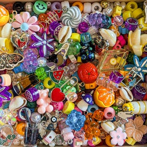 Op de afbeelding: Een grote verzameling kleurrijke plastic en glazen kralen, bedels en hangers. De bedels omvatten een verscheidenheid aan vormen, zoals harten, sterren, vlinders en bloemen. De kralen zijn in verschillende kleuren, waaronder rood, blauw, groen, geel en paars.