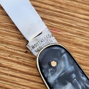 May include: Close-up of a vintage pocket knife with a silver blade and a black and white marbled handle. The blade is engraved with the text "ROBI. KLAAS SOLINGEN". The handle has a gold-colored rivet.