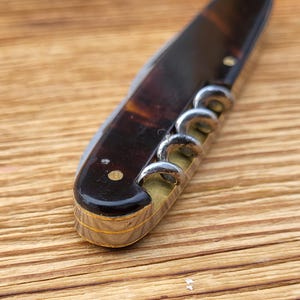 May include: A vintage corkscrew with a tortoiseshell handle and a silver-colored worm. The handle has a dark brown and amber pattern. The corkscrew is lying on a wooden surface.