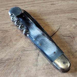 May include: A vintage pocket knife with a white and black handle and a corkscrew. The knife has a silver blade and a silver cap.
