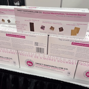 May include: A pink and white box of Sweet Marshmallow Co. Gourmet Marshmallows. The box features a photo of marshmallows and graham crackers. The text on the box reads "Gourmet Marshmallows/Guimauves Gourmet" and "Gourmet S'mores Kit".