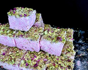 May include: A stack of pink and white squares covered in green pistachio nuts and pink rose petals. The squares are arranged in a pyramid shape on a black surface.