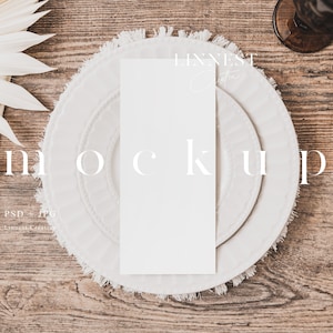May include: A white card mockup on a white plate with a fringed edge. The plate is on a wooden table with a white napkin and a decorative fan.