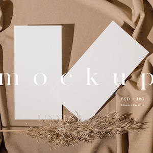 Pode incluir: Dois cartões brancos em branco sobre um fundo de tecido marrom com um caule de grama seca. O texto "mockup" está escrito em branco nos cartões. O texto "PSD + JPG" e "Linness Creative" está escrito em branco na parte inferior da imagem.
