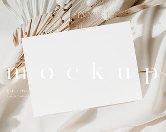 May include: A white card mockup with the word "mockup" printed in white on the card. The card is lying on a white fabric with dried palm leaves and white fluffy flowers.