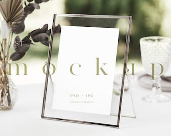 May include: A clear acrylic picture frame with a white card inside. The frame is standing on a white table with a vase of dried flowers and a glass on the table.