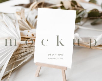 May include: A white card mockup on a small wooden easel, surrounded by dried palm leaves and other natural elements. The card is blank and ready for your design. PSD + JPG. Linnest Creative.