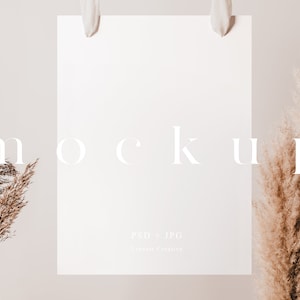 May include: A white poster mockup hanging from a wooden frame with white fabric ties. The poster is blank and has the text "MOCKUP" and "PSD + JPG | Lemon Creative" printed on it. The poster is surrounded by dried pampas grass.