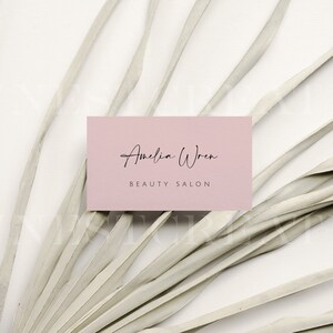 May include: A pink business card with the text "Amelia Wren Beauty Salon" in black script font. The card is lying on a white surface with a dried palm frond.