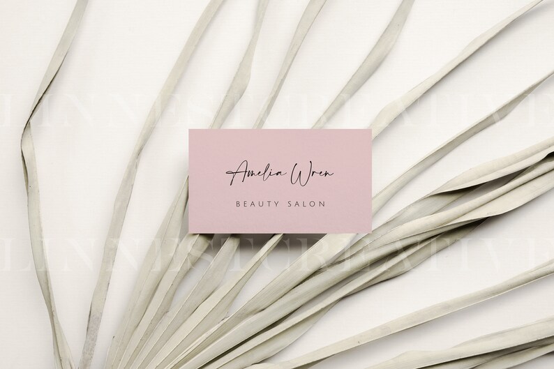 May include: A pink business card with the text "Amelia Wren Beauty Salon" in black script font. The card is lying on a white surface with a dried palm frond.