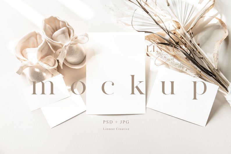 May include: A mockup featuring three blank cards with the word "mockup" printed in brown text. The cards are arranged on a white surface with a bouquet of dried palm leaves and two pairs of beige baby booties.