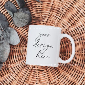 K&ouml;nnte beinhalten: Wei&szlig;e Keramiktasse mit einer schwarzen Aufschrift "your design here" auf einem geflochtenen Weidenhintergrund.