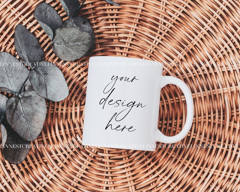 K&ouml;nnte beinhalten: Wei&szlig;e Keramiktasse mit einer schwarzen Aufschrift "your design here" auf einem geflochtenen Weidenhintergrund.
