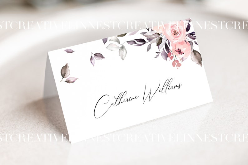 May include: A white place card with a floral design in shades of pink and gray. The name "Catherine Williams" is written in cursive on the card.