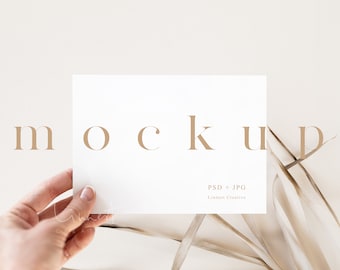 May include: A white card mockup with the word "mockup" in brown text. The card is being held by a hand and is surrounded by dried beige grass.
