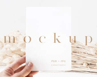 May include: A white card mockup with the text "PSD + JPG" and "Linnest Creative" in brown text. The card is being held by a hand against a background of dried beige grass.
