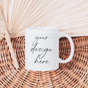 K&ouml;nnte beinhalten: Wei&szlig;e Keramiktasse mit einer schwarzen Aufschrift "your design here" auf einer geflochtenen Weidenoberfl&auml;che mit einem getrockneten F&auml;cher aus Palmblatt.