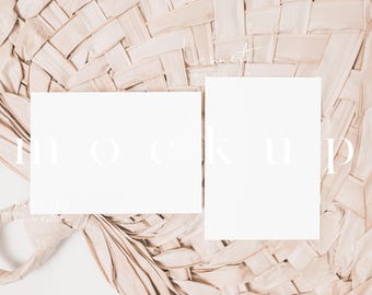 May include: Two blank white cards on a woven straw background. The cards are arranged side by side and the text "MOCKUP" is written in white on the background.