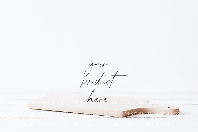 May include: A light brown wooden cutting board with a handle, sitting on a white wooden surface. The text "your product here" is written in black cursive on the board.