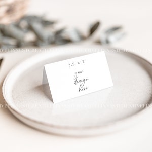 May include: A white place card with the text "3.5 x 2" and "your design here" on a white plate with gold silverware.