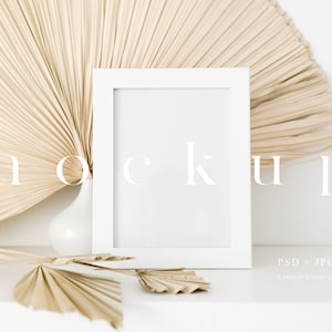 May include: White picture frame mockup with a beige dried palm leaf fan behind it. The frame is sitting on a white surface with a smaller dried palm leaf fan in front of it.