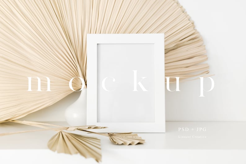 May include: White picture frame mockup with a beige dried palm leaf fan behind it. The frame is sitting on a white surface with a smaller dried palm leaf fan in front of it.